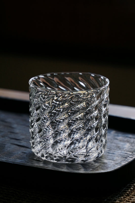 Straight-sided clear glass cup covered in a fine diagonal scale pattern, on a dark tray.