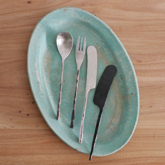 A spoon, fork and two knives with slim handles laid across a pale turquoise oval plate.