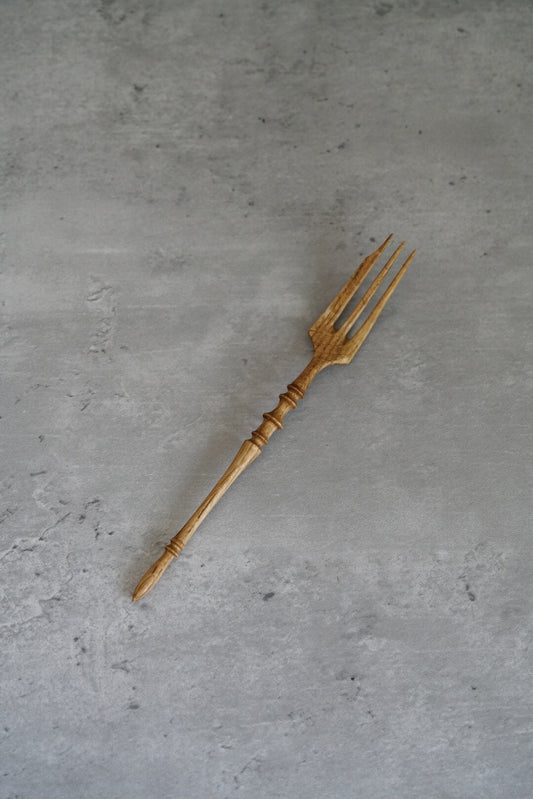 Carved wooden fork with three slim tines and a turned handle, on grey concrete.