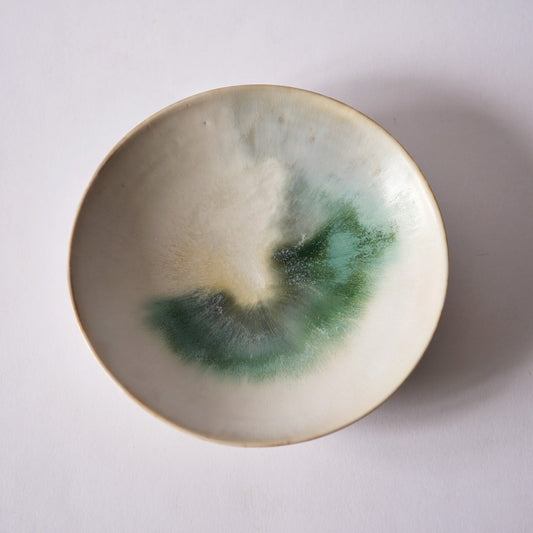 Cream plate seen from above, green and brown glaze pooling in a soft crescent across its centre.