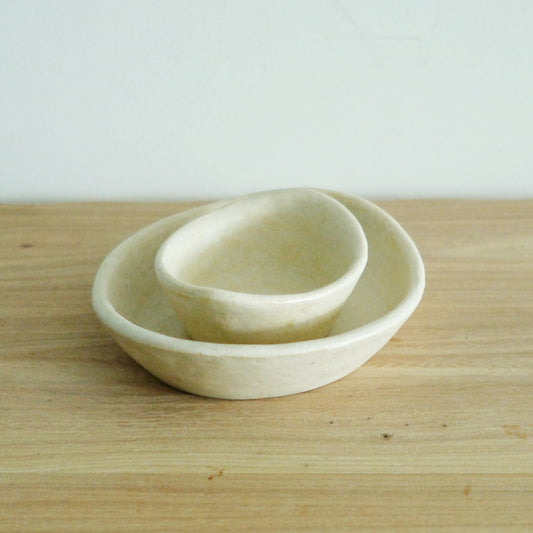 Two nested oval bowls in soft cream and pale yellow with rough unglazed outsides, on wood.