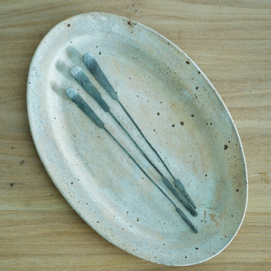 Three long slim tea spoons with pale blue tips, laid diagonally across a speckled oval plate.