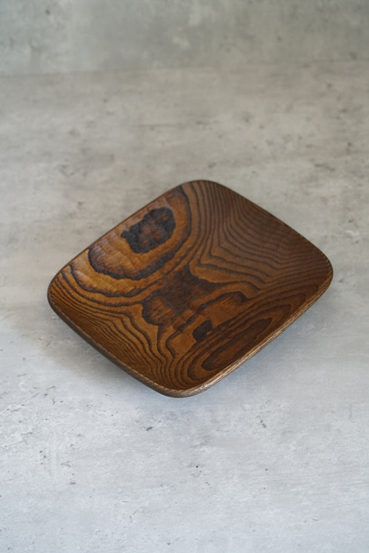 Square wooden dish in warm amber lacquer, its bold grain swirling across the surface.