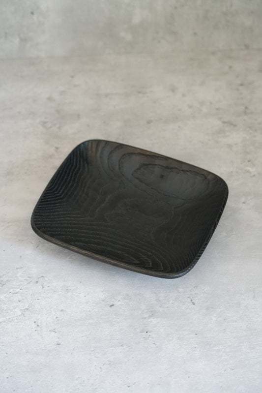 Square wooden dish in matte black lacquer, its grain running in fine straight lines.