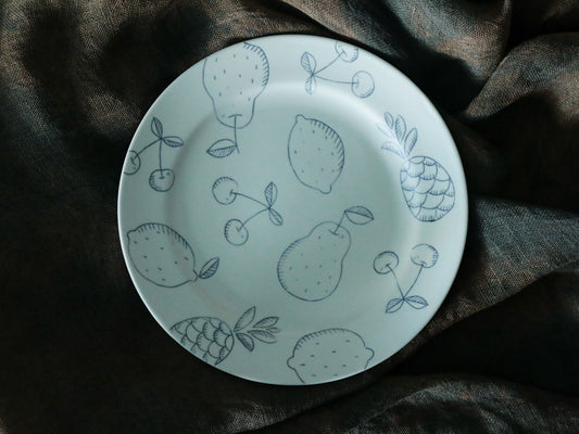 Pale porcelain plate drawn in fine grey line-work with lemons, pears, cherries and a pineapple.