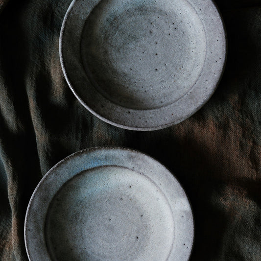 Two speckled grey-blue plates with raised rims, seen from above on dark brown cloth.