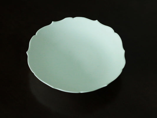 Shallow celadon plate with a softly scalloped edge, glowing pale green against black.