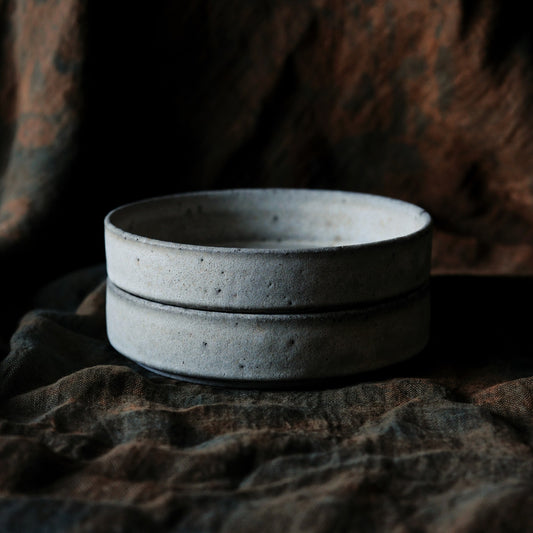Two wide flat speckled grey bowls stacked one on the other, on dark brown cloth.