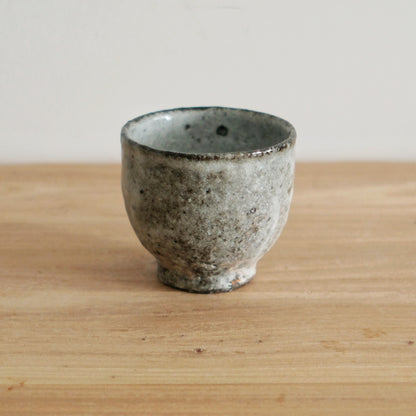 Footed wood-fired cup in grey and white with a rough speckled surface, on a light wooden table.