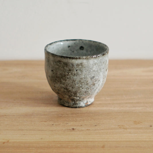Footed wood-fired cup in grey and white with a rough speckled surface, on a light wooden table.
