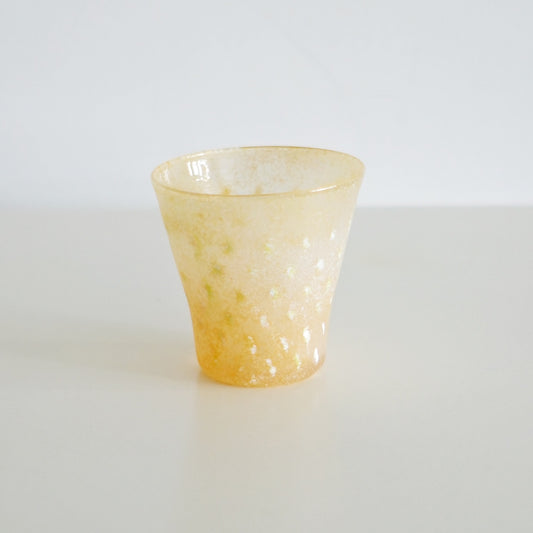Flared glass cup in warm amber-yellow with a mottled, frosted surface, on a pale table.