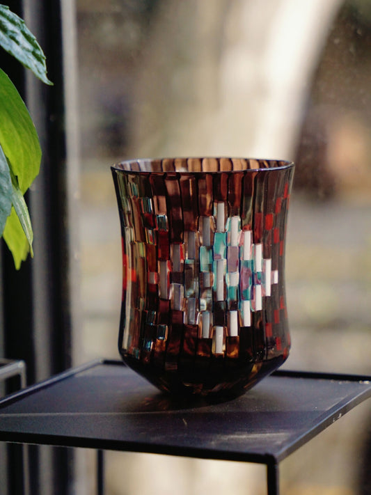 Tall glass built from a mosaic of dark red, white and blue tiles, standing on a black stand outdoors.