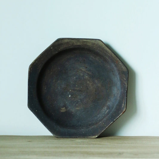 Octagonal plate in dark iron grey with a rough matte surface, standing upright on a wooden shelf.