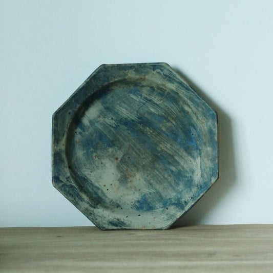 Octagonal plate washed in dusty blue and grey with brushed streaks, standing on a wooden shelf.