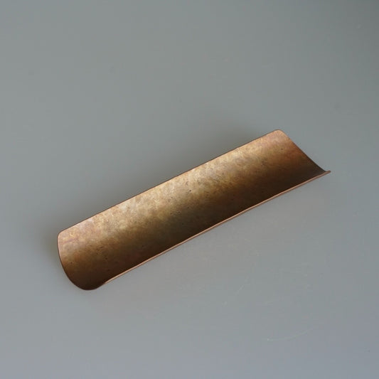 Long narrow brass tea scoop rolled into a shallow channel, its surface warmly hammered.