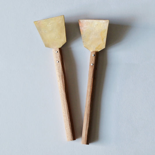 Two small brass spatulas with slim wooden handles, laid side by side on a pale blue ground.