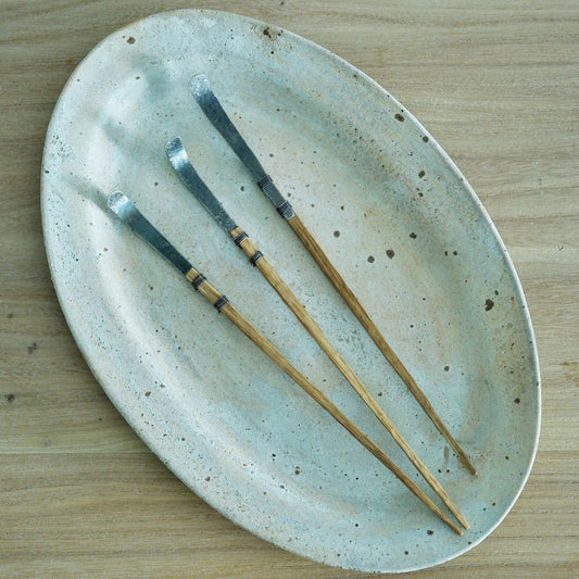 Four long tea scoops with slim wooden handles and metal bowls, laid across a speckled oval plate.