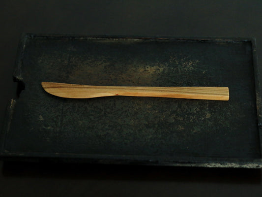 Slender carved wooden spreading knife with a tapered blade, on a dark metal tray.