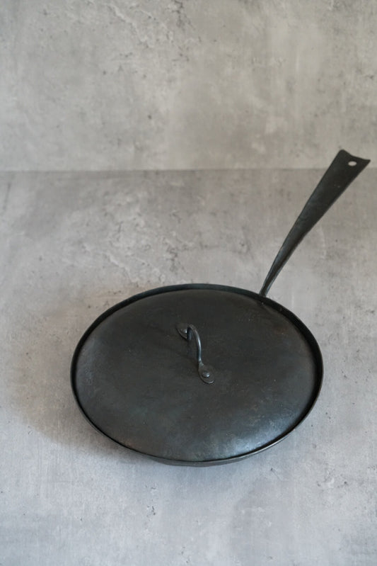 Round black iron frying pan with a fitted lid and a long flat handle, on grey concrete.
