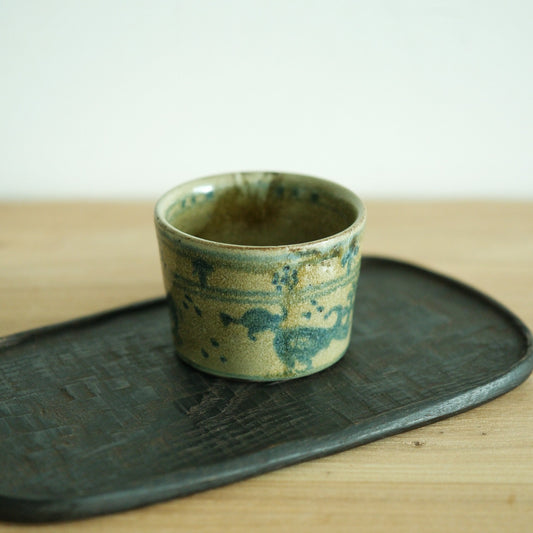 Straight-sided cream cup with pale green and blue glaze pooling around its base, on a dark board.