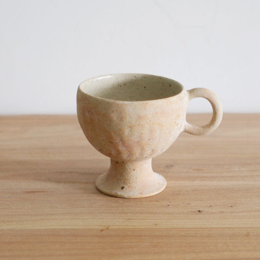 Cream stemmed cup with a small ring handle and a sandy speckled surface, on a wooden table.