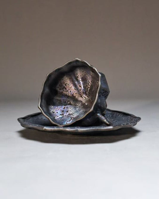 Flower-shaped cup and saucer in dark iridescent glaze, the petals catching bronze and violet highlights.