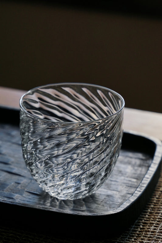 Round clear glass cup with a twisted scale-like texture running around it, on a dark tray.