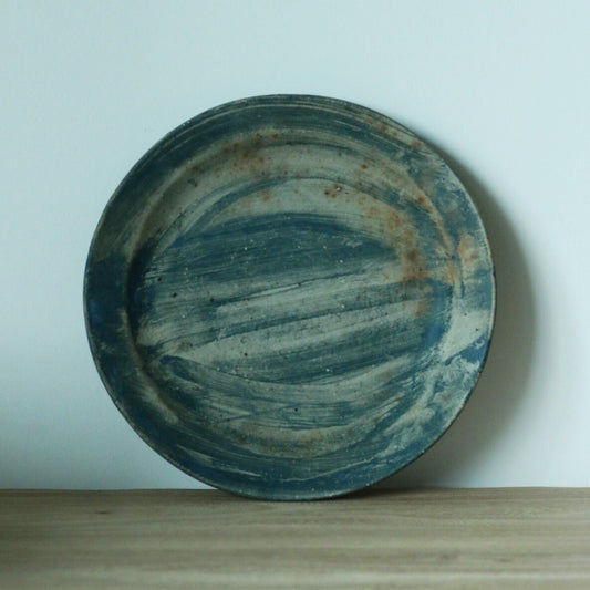 Round plate brushed in slate blue over warm brown, standing upright on a wooden shelf.