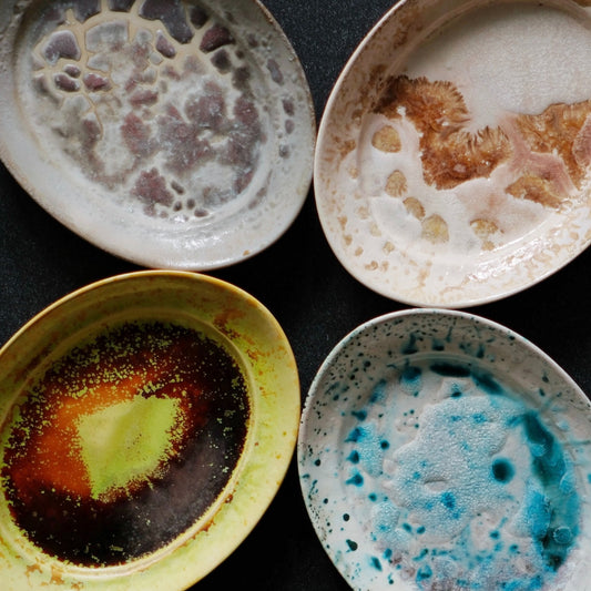 Four plates with flowing glazes in amber, turquoise, rust and speckled white, arranged on black.
