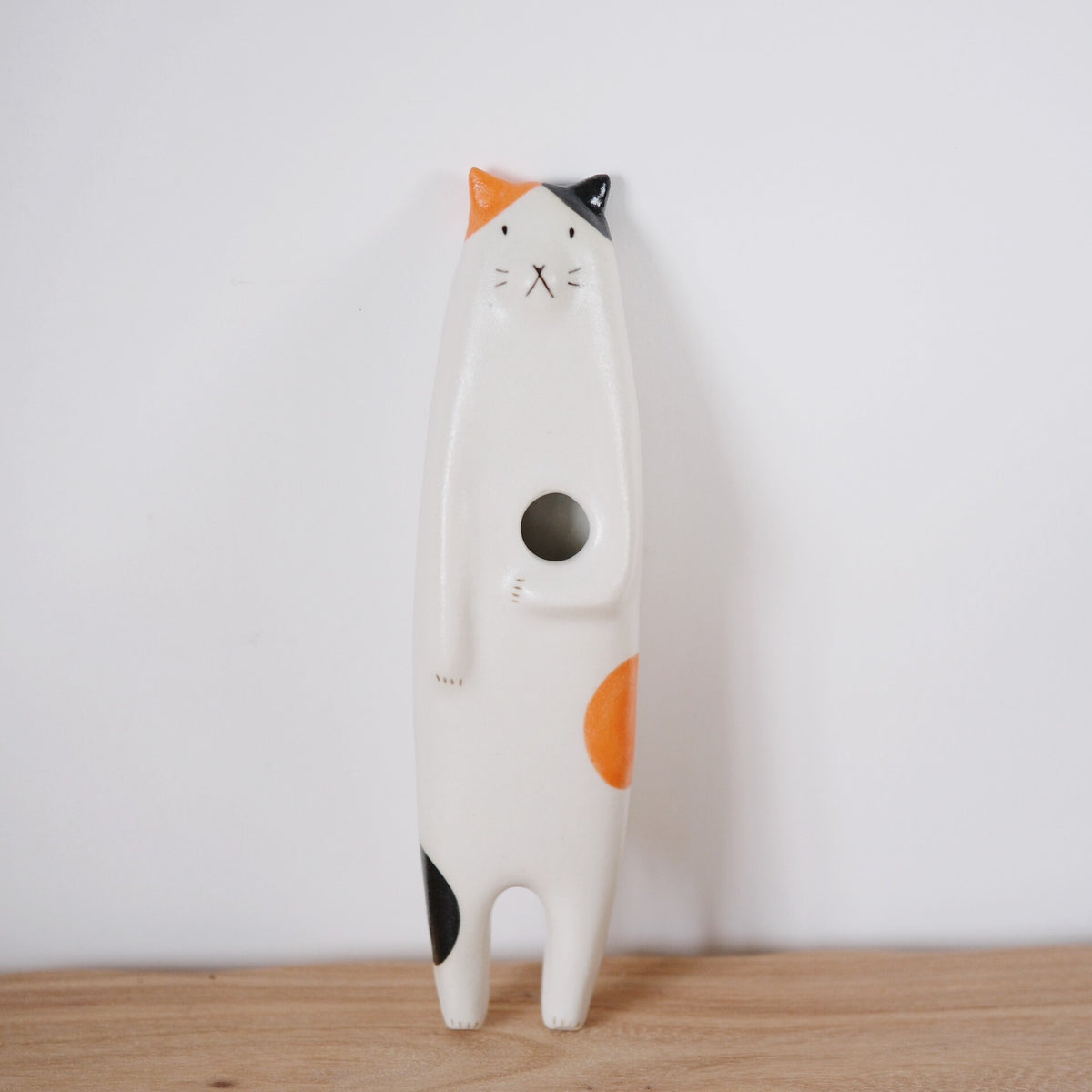 Tall standing ceramic cat in white with orange and black patches and a round opening in its belly.