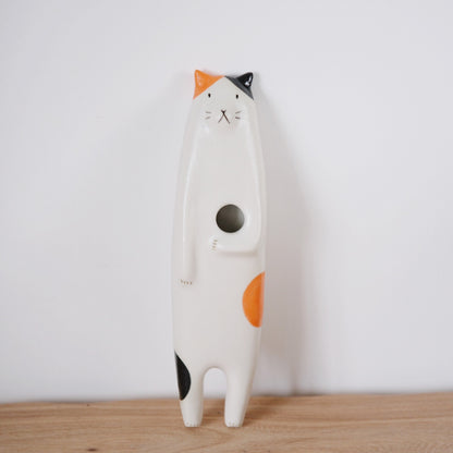 Tall standing ceramic cat in white with orange and black patches and a round opening in its belly.
