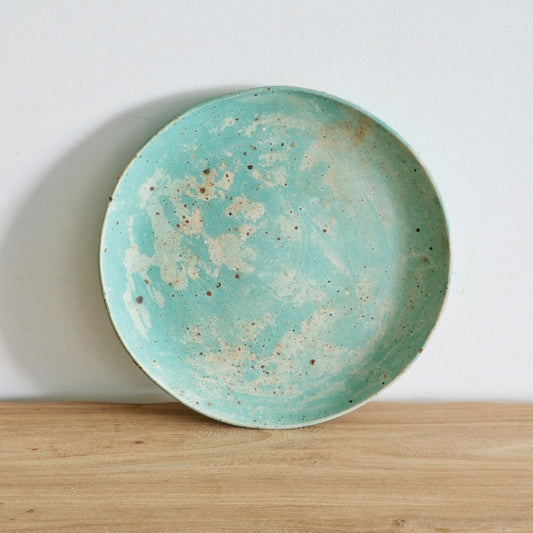 Round plate in pale turquoise clouded with cream, standing upright on a wooden shelf.