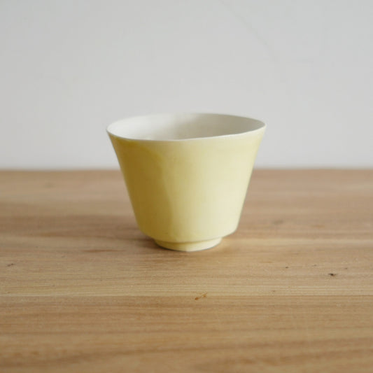 Small footed cup in soft primrose yellow with a thin flared rim, on a light wooden table.