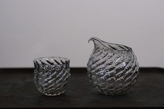 A small ribbed glass cup and a matching pouring bowl in pale smoky grey, side by side on white.