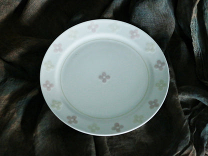 White porcelain plate scattered with small pale pink four-leaf clover motifs, on dark cloth.
