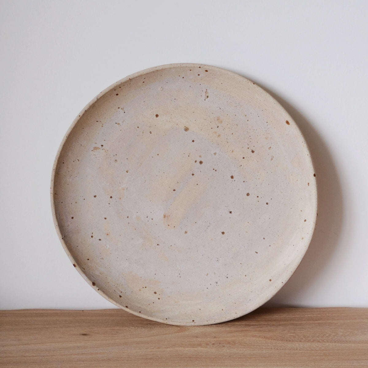 Large round plate in speckled cream with a faint blush of pink, standing on a wooden shelf.