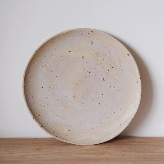 Large round plate in speckled cream with a faint blush of pink, standing on a wooden shelf.