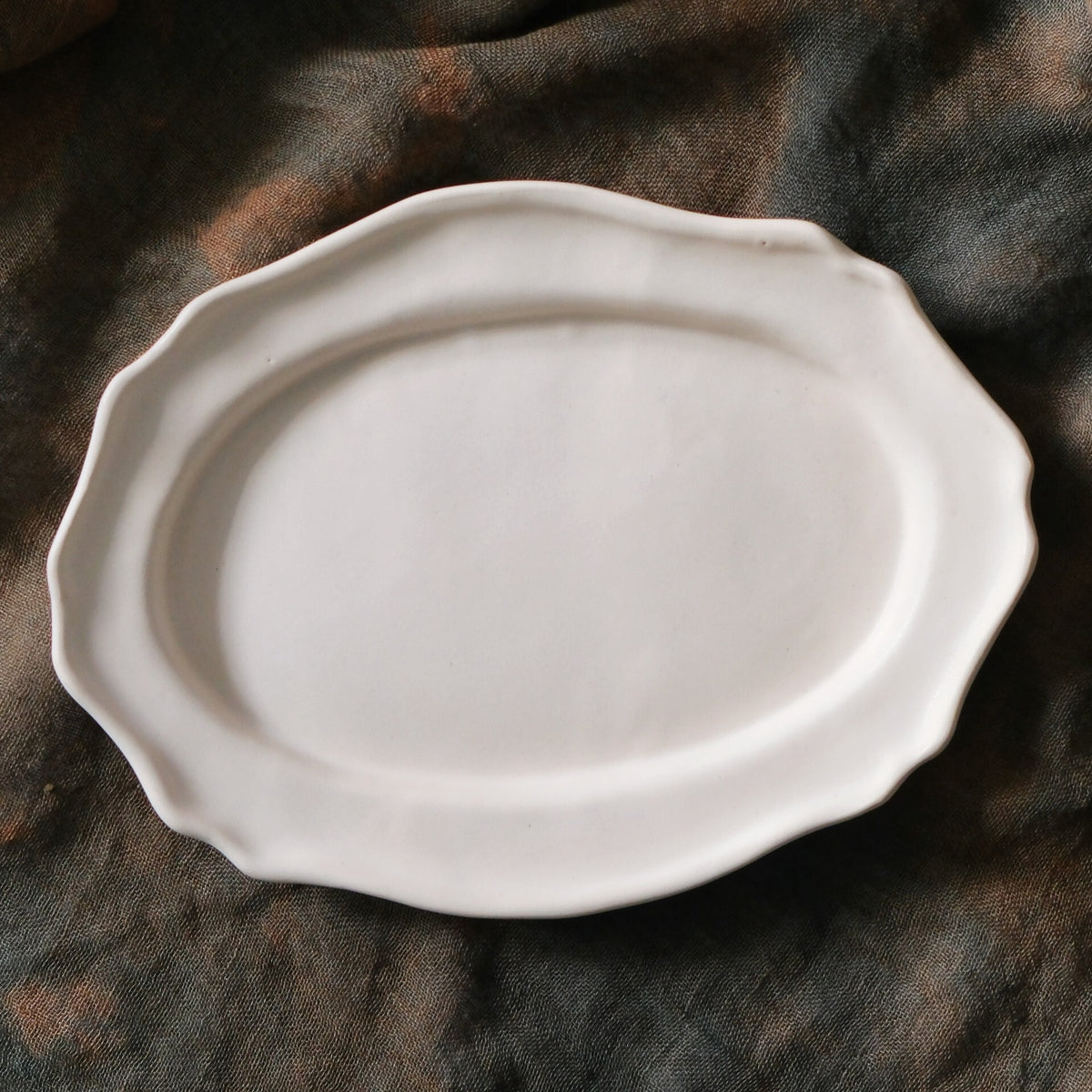 Oval plate in soft chalky white with a gently scalloped rim, on dark brown cloth.