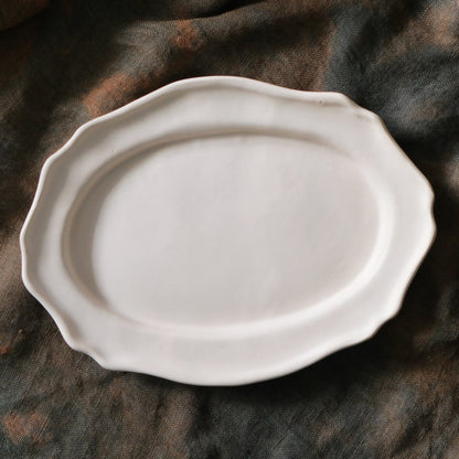 Oval plate in soft chalky white with a gently scalloped rim, on dark brown cloth.