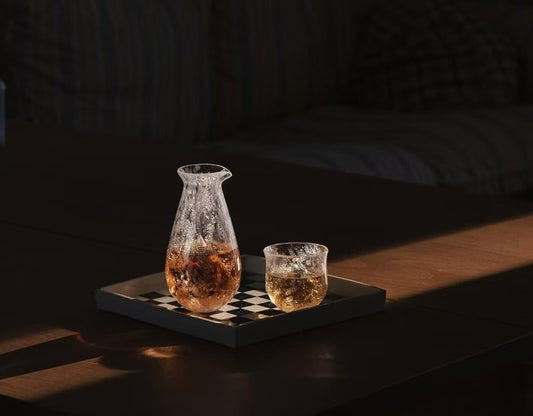 Slender clear glass sake carafe and a small matching glass, both holding amber liquid, on a dark chequered tray.