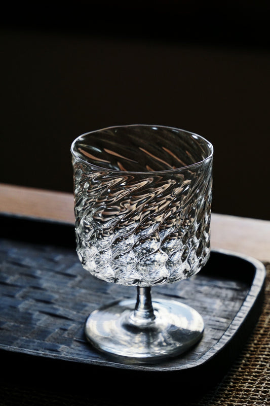 Tall clear glass cup on a short stem, its body worked in a twisted scale pattern, on a dark tray.