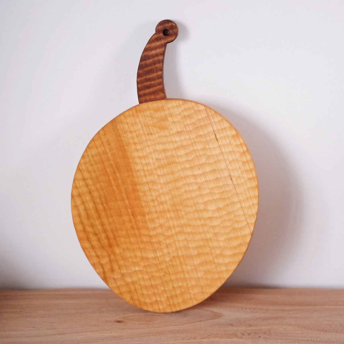 Round wooden bread board with a rippled grain and a short curved handle, leaning against a wall.