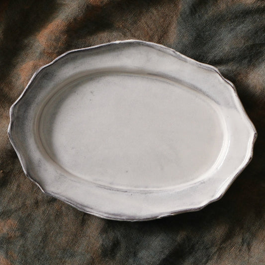 Oval plate in chalky white with a waved scalloped rim and soft grey shading, on brown cloth.
