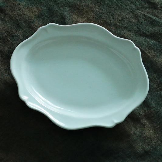 Oval celadon plate in pale blue-green with a softly waved scalloped rim, on dark cloth.