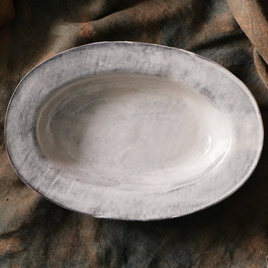 Oval plate in chalky off-white with a deep well and softly brushed grey edges, on brown cloth.