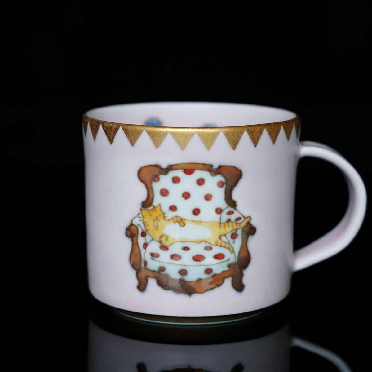 White porcelain mug with a gold triangle band at the rim, painted with a cat curled on a red-spotted armchair.
