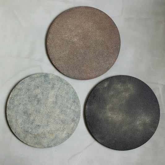 Three round matte serving discs laid on cloth, one warm brown, one pale blue-grey and one black.
