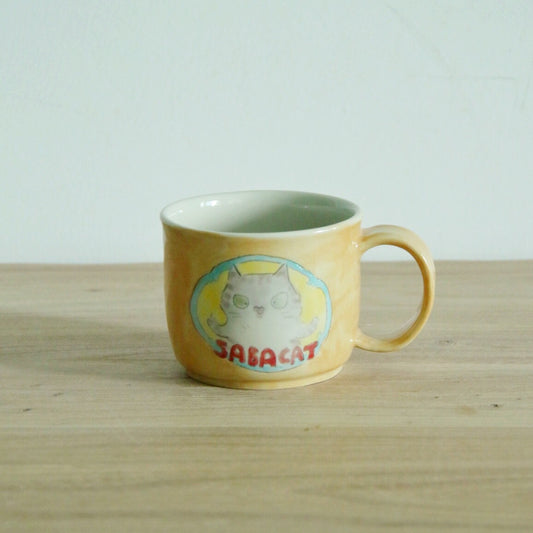 Soft yellow mug painted with a round cat inside a blue oval, with a matching yellow handle.