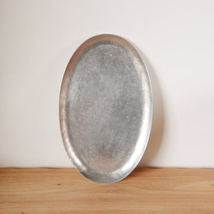 Plain oval aluminium tray with a softly brushed silver surface, standing upright on a wooden shelf.