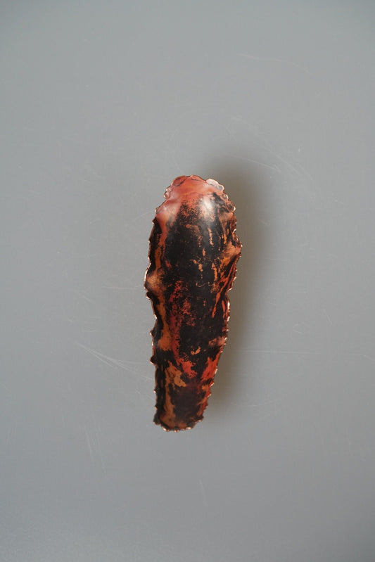 Slim brass tea scoop finished in fiery red and black, mounted against a pale wall.